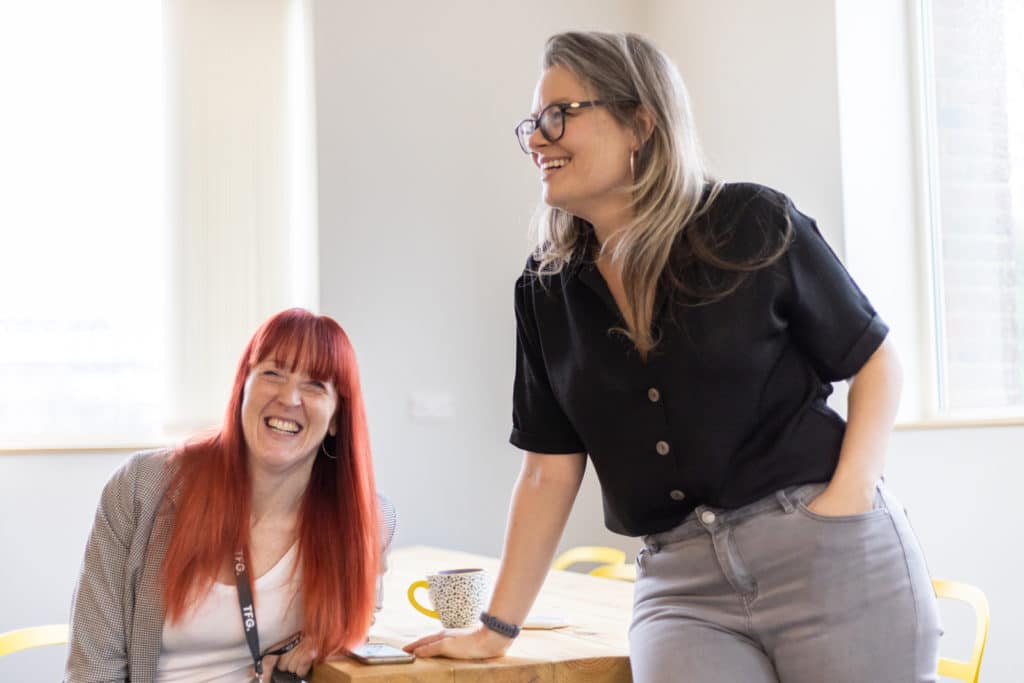 Natalie Welch and Polly Buckland owners of Hampshire Marketing Agency The Typeface Group. Natalie is sitting at the table while Polly is perched on the table. Both laughing.