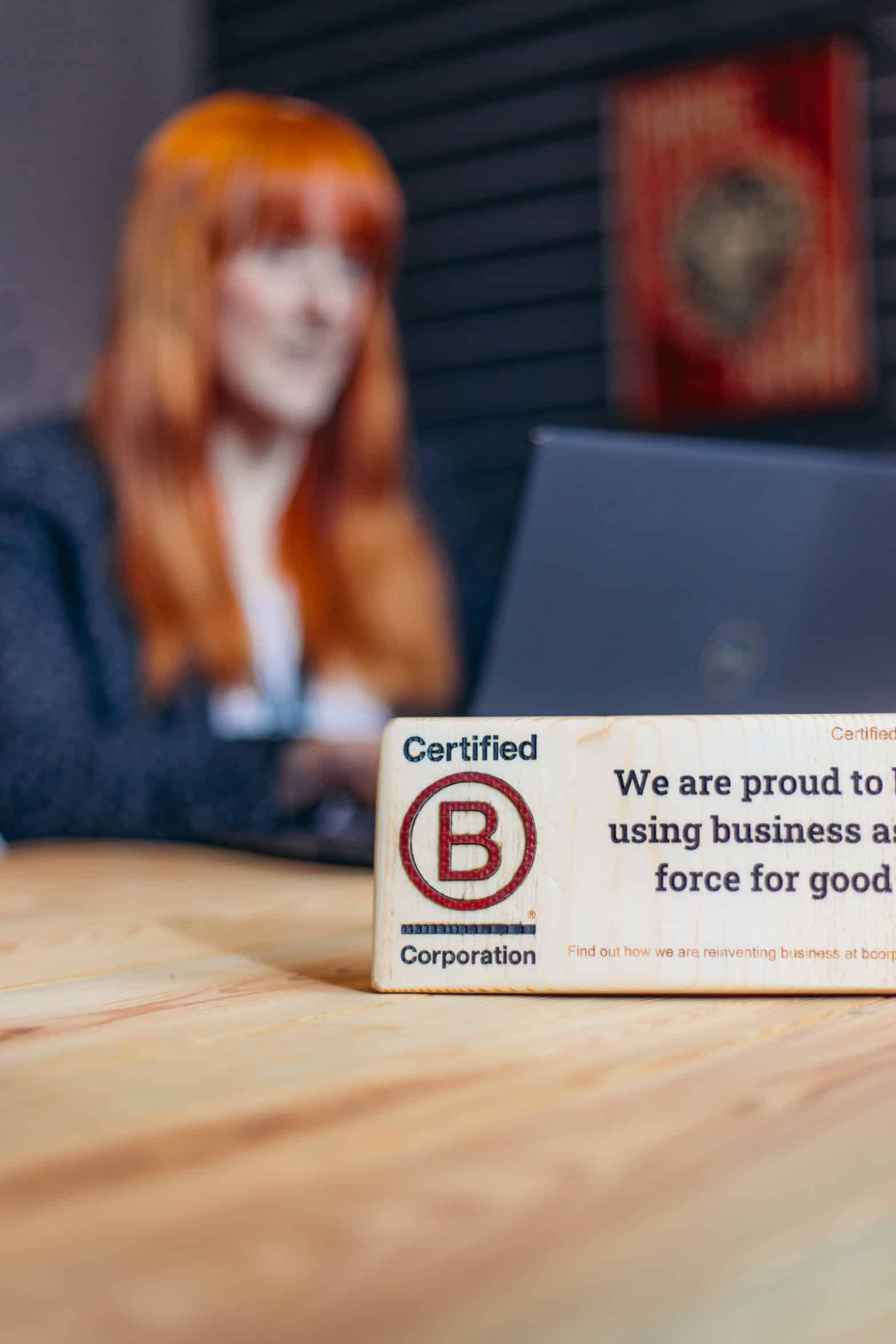 Natalie in the background typing on a laptop with a wooden plaque saying "Certified B Corporation. Certified in 2021 We are proud to be a business as a force for good. Find out how we are reinventing business at bcorporation.co.uk